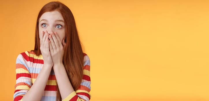 Shocked upset redhead female witness terrible accident gasping cover mouth palms astonished staring left frightened express empathy worry standing orange background panicking. Copy space
