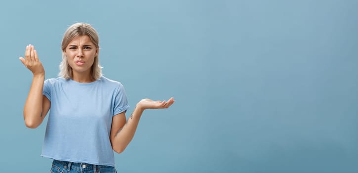 Wtf you saying. Portrait of pissed and confused stylish girlfriend with blond hair raising palm high and aside with perplexed expression being questioned hearing stupid nonsense over blue wall.