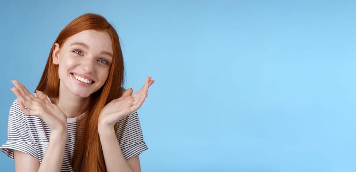 Silly cute sincere young european ginger girl blue eyes shrugging hold hands doubtful spread near face tilting head apologizing flirty say sorry late making lame excuses, standing blue background.