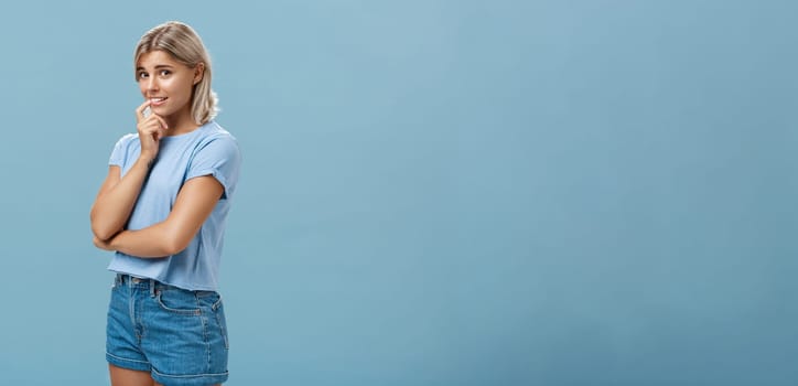 Studio shot of unconfident silly and insecure cute feminine girl with blond hair standing half-turned over blue background holding finger on lip while being shy ask question smiling hesitating. Emotions concept