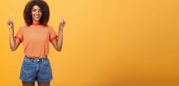Looking only up and forward. Optimistic ambitious stylish dark-skinned female student in striped cool t-shirt and shorts raising hands pointing upwards and smiling friendly over orange wall.