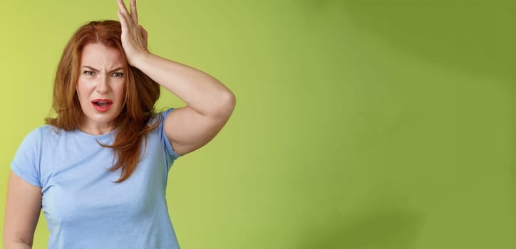 Ouch you kick my head. Frustrated concerned upset middle-aged redhead woman look shocked distressed touch temple complain kid shoot ball her face stand disappointed green background. Copy space