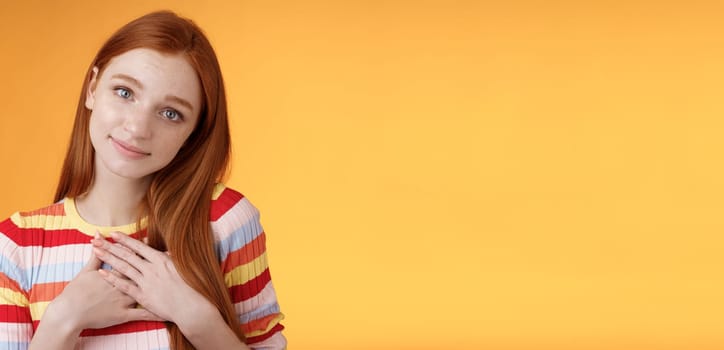 Lovely silly cute redhead young european girl tilting head look gently smiling tender camera press palms heart thankful receive nice gift gazing gratitude happiness, standing orange background.