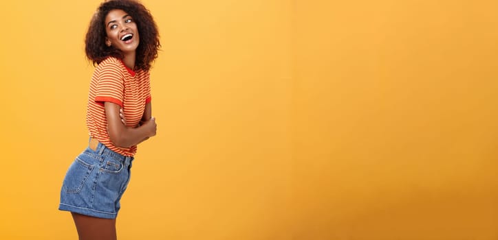 Hey wanna hang out. Sensual and flirty carefree stylish dark-skinned woman with curly hair standing in profile turning left and posing with happy cute expression smiling and seducing over orange wall.