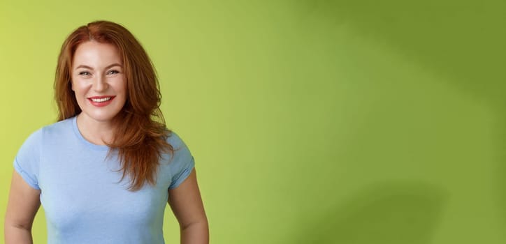 Cheerful kind happy redhead middle-aged mother look caring delighted smiling broadly gaze admiration joy stand blue t-shirt green background amused enthusiastic expression. Copy space