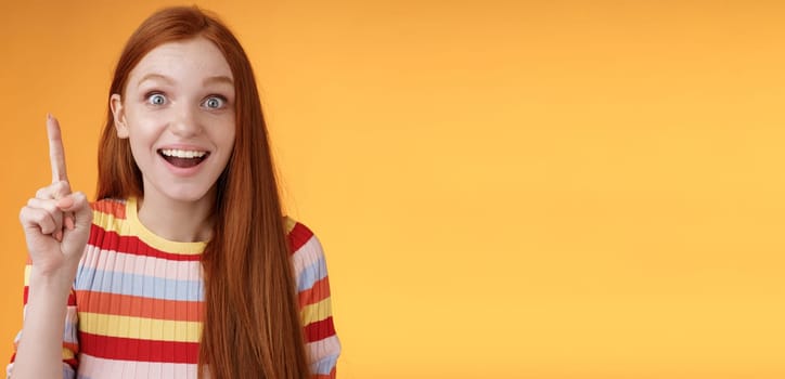 Excited happy cheerful young redhead female student got idea think answer raising index finger eureka gesture smiling thrilled wide eyes saying plan have excellent suggestion, orange background.