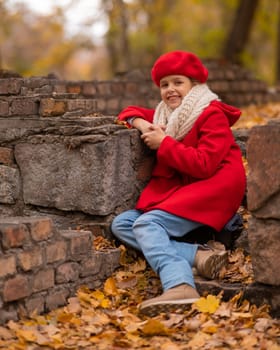 Smiling caucasian girl in a red coat and beret sits on a brick wall on a walk in autumn