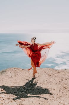 Side view a Young beautiful sensual woman in a red long dress posing on a rock high above the sea during sunrise. Girl on the nature on blue sky background. Fashion photo.