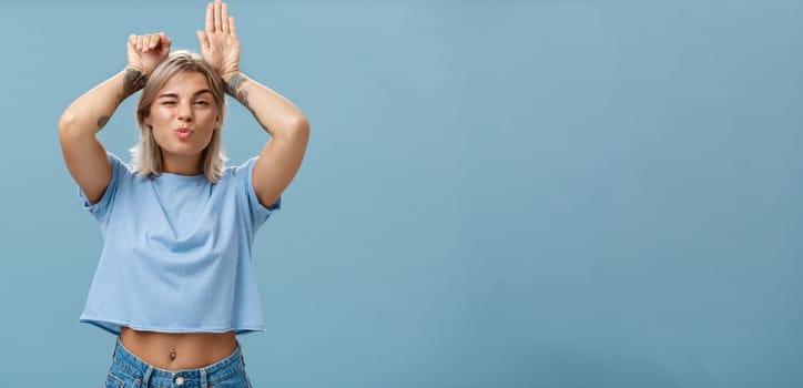 Lifestyle. Flirty attractive and stylish cool girlfriend with tattooed arms and pierced belly winking folding lips in air kiss showing bunny ears with palms on head standing over blue background playful.