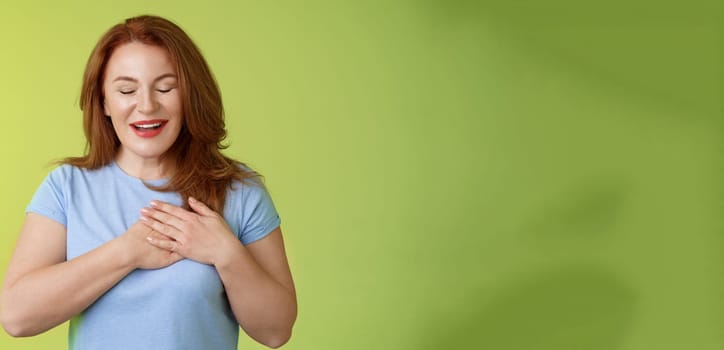 Fascinated cute redhead passionate middle-aged woman sighing lovely touch heart close eyes smiling delighted express admiration temptation feeling appreciation grateful emotions green background.