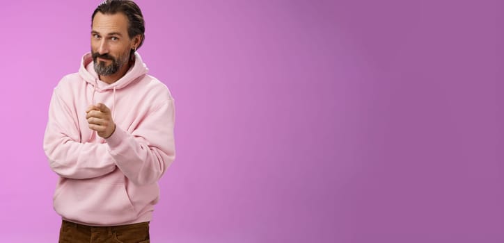 Confident handsome charismatic adult bearded man in stylish pink hoodie inviting you join company smiling self-assured indicating picking make decision chosing us, purple background.