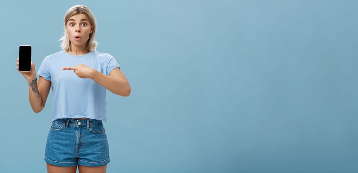 Wow look what found online. Portrait of impressed speechless good-looking young sportswoman in t-shirt and denim shorts saying wow folding lips showing smartphone and pointing at screen over blue wall. Technology concept