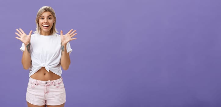 Lifestyle. Indoor shot of delighted and amused attractive stylish urban female with blond hair in stylish white t-shirt and summer shorts raising palms from surprise and amazement standing over purple background.