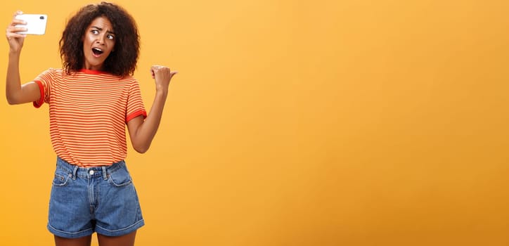 Woman recording video blog pointing at strange object behind her. Portrait of concerned and curious stylish famous internet star holding smartphone talking in cellphone camera over orange background. Technology concept