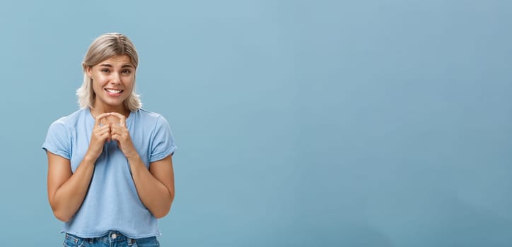 Silly cute and worried blonde female friend with short haircut steepling index fingers and smiling with guilty sorry smile frowning while waiting for answer begging for help or favor over blue wall. Copy space