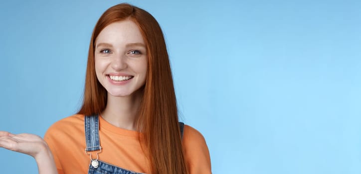 Proud good-looking confident redhead girl present awesome product hold object palm raise hand blank blue copy space smiling delighted recommend cool link, standing studio background helpful.
