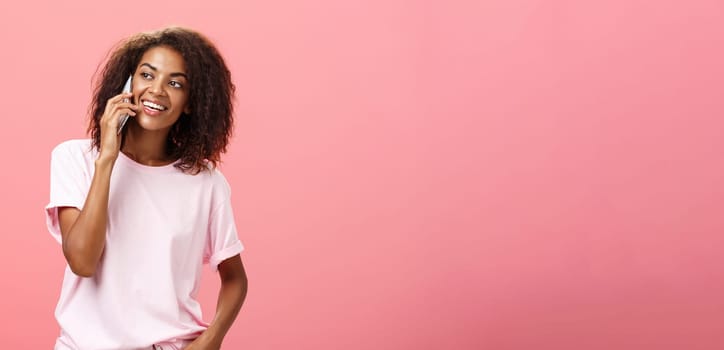 Girl calling friend to meet up. Portrait of charming friendly and outgoing african american young woman with afro hairstyle holding smartphone near ear while talking looking left curiously. Copy space