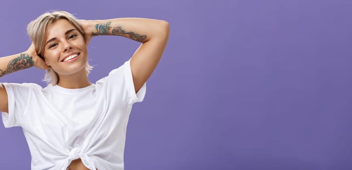 Lifestyle. Close-up shot of good-looking stylish and relaxed blond woman with tattoos on arms holding hands behind head smiling with carefree pleased look enjoying weekends over purple background.