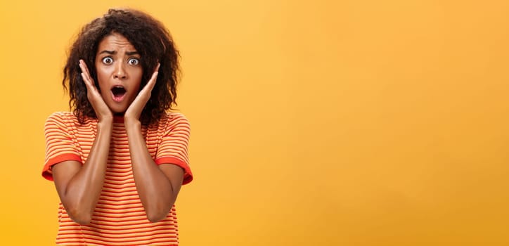 Waist-up shot of shocked concerned panicking african-american woman with afro hairstyle in trendy t-shirt gasping holding palms on face from surprise posing troubled against orange wall. Lifestyle.