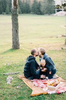 Mom kisses dad while sitting on her knees with a little boy on her lap on a blanket in the park. High quality photo