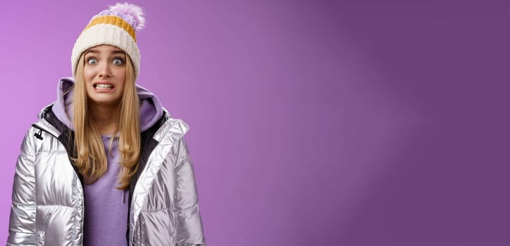 Awkward worried cute timid blond girl in silver jacket hoodie winter hat clench teeth popping eyes camera ooops make mistake standing nervous someone notice, purple background. Copy space