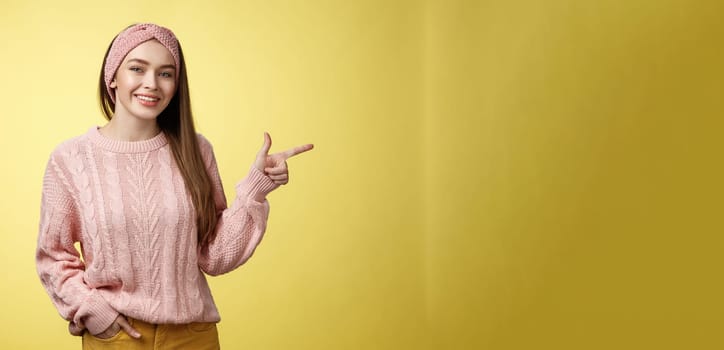 Lifestyle, people, pointing concept. Attractive young girl with long straight hair wearing sweater pointing aside using fore finger, indicating copy space smiling cheerful, positive over yellow wall.