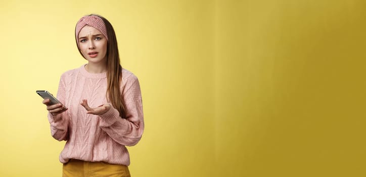 Puzzled confused upset questioned young cute girl in headband knitted sweater frowning clueless raising hand in dismay holding smartphone, cannot understand what happened cellphone over yellow wall.