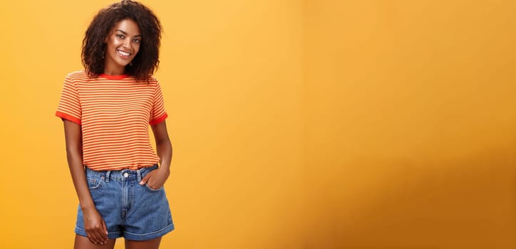 Ready to travel world. Energetic confident and attractive dark-skinned woman with curly hair holding hand in pocket of denim shorts smiling joyfully posing over orange background carefree and friendly.