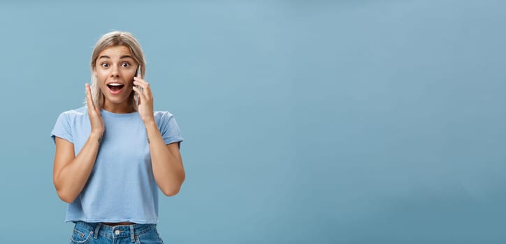 Pleased to hear best friend calling from abroad. Portrait of surprised pleased charming blonde female in trendy t-shirt opening mouth from amazement holding smartphone near ear and hand on face. Technology concept