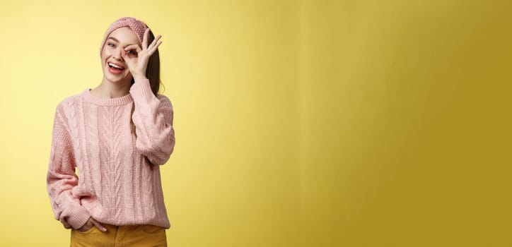 Cheerful girl saying ok. Attractive stylish shoolgirl in knitted sweater relaxed feeling upbeat, sharing happiness and joy, showing okay sign on eye smiling optimistic, entertained over yellow wall.