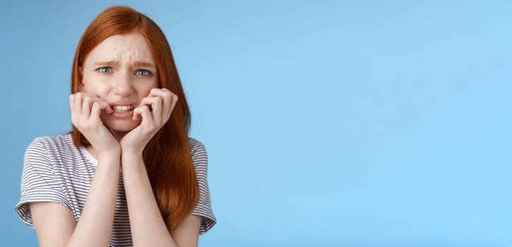 Worried uncomfortable scared young panicking redhead young girl feeling pressure distressed frowning squinting frightened biting fingernails trembling fear, standing blue background.