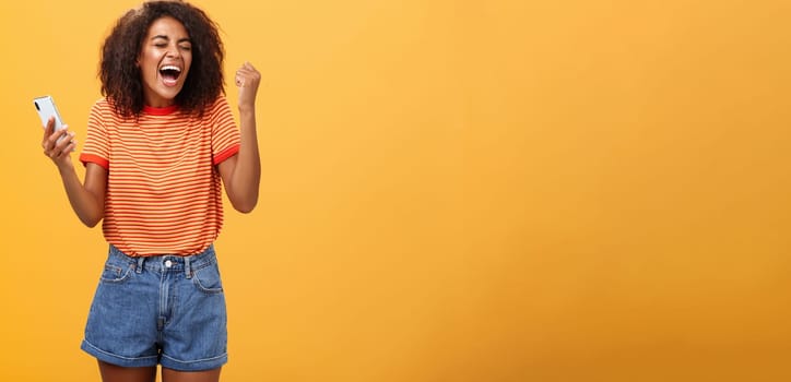 Portrait of ambitious happy young african american girl yelling from happiness and triumph clenching fist in joy and celebration feeling excited and relieved holding smartphone over orange wall. Lifestyle.