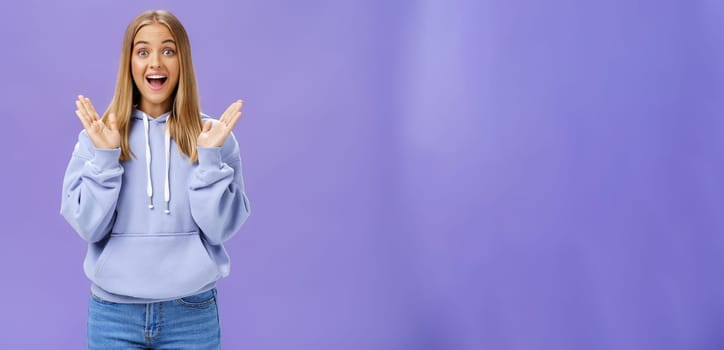 Portrait of amused excited and entertained happy charming woman with fair hair in trendy hoodie clasping hands from delight and surprise smiling broadly joyfully posing against purple background. Lifestyle.