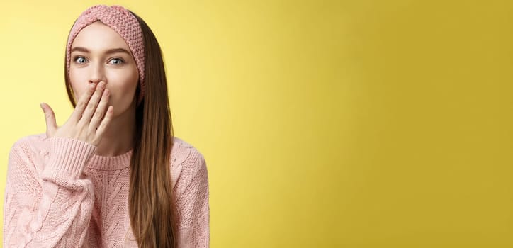 Talkative young cute office worker wearing sweater, knitted headband holding palm on mouth surprised and amazed, learning interesting shocking rumor, gossiping entertained over yellow wall.