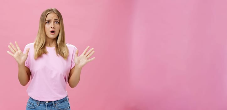 Woman looking nervous explaining with panicking gestures she not involved frowning opening mouth and gasping feeling concerned and worried waving hands over chest posing against pink wall. Copy space