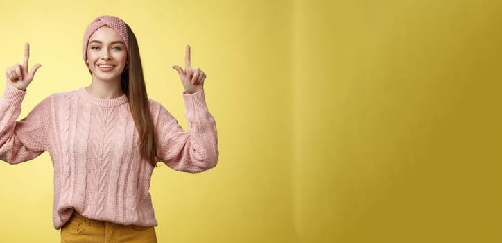 Easygoing beautiful young female student in knitted sweater, headband pointing up, promoting advertisement, smiling happily, feeling positive posing in good mood, grinning at camera over yellow wall.