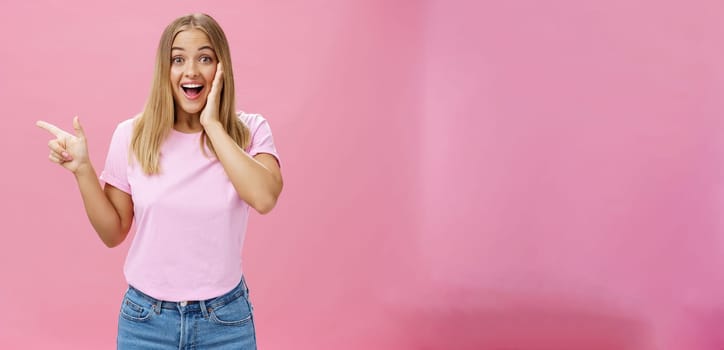 Portrait of surprised enthusiastic and charismatic tanned cute woman with fair straight hair touching cheek from joy and amazement pointing left delighted and amused over pink background. Copy space