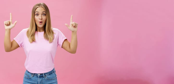 Amused and impressed attractive common european woman with fair hair and tanned skin raising index fingers pointing up amazed folding lips from interest and astonishment over pink background. Copy space