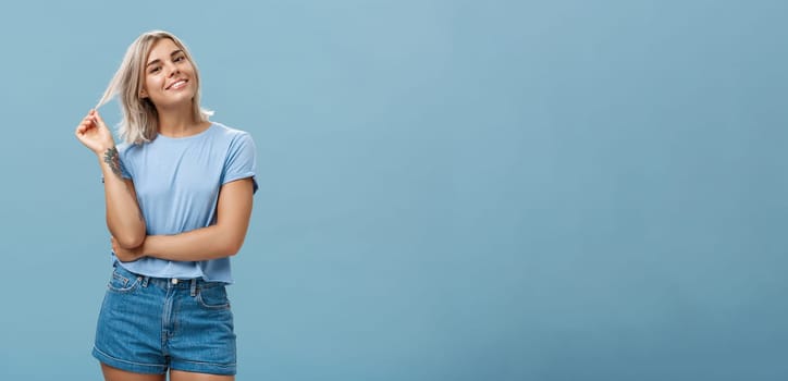 Studio shot of relaxed and chill popular attractive woman with blond hair and tattoos playing with strand tilting head and smiling as if listening boyfriend or flirting standing amused over blue wall. Body language and emotions concept