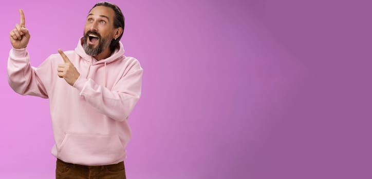 Fashionable good-looking fascinated mature bearded man grey hair look astonished pointing gazing upper left corner see miracle incredible breathtaking magnificanent view, standing purple background.