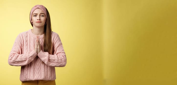 Deliberately sad girl in sweater pressing palms together in pray pouting praying for help, asking favor, begging friend lend cute dress for date, smiling silly, want something over yellow background.
