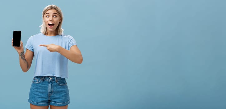 Studio shot of amazed and impressed charming european blonde in casual clothes gasping charmed at camera while showing smartphone screen pointing at device with finger over blue background. Technology and advertisement concept