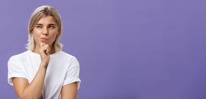 Waist-up shot of smart creative young ambitious woman with blond haircut and tanned skin squinting and smirking gazing left while holding hand on chin thinking making up plan or decision. Copy space