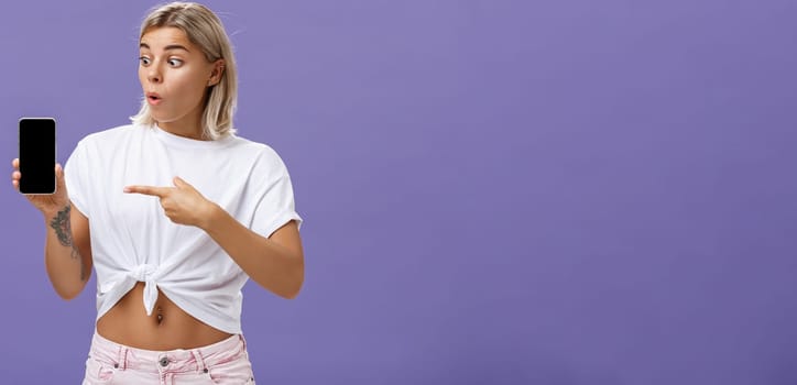 Waist-up shot of impressed and delighted gorgeous european female with blond hair and tattoos looking and pointing at awesome brand new smartphone standing over purple background. Technology concept