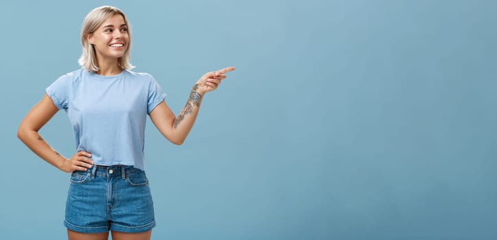 Bossy confident female manager with tattoo on arm holding hand on waist pointing and gazing right with pleased relaxed look giving directions to employees posing over blue background. Copy space