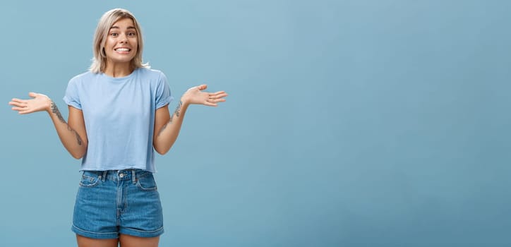 Oops I do not know. Silly cute european girl with fair hair and cool tattoos shrugging with palms spread aside in no idea gesture smiling with sorry look being unaware and clueless over blue wall.