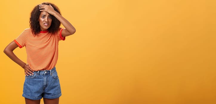 Shit I am in trouble. Concerned anxious cute dark-skinned woman in trendy t-shirt and shorts clenching teeth with guilty look frowning punching forehead having trouble or problem over orange wall.
