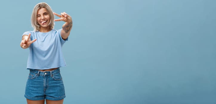 Beautiful tattooed girl enjoying weekends sticking out tongue joyfully winking at camera and smiling showing peace or victory gesture with pulled arms feeling happy posing over blue background. Copy space