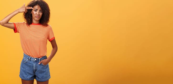 Portrait of displeased gloomy and sad annoyed dark-skinned female with curly hairstyle showing gun with hand near temple frowning wanting to kill herself from exhaust and boredom over orange wall. Lifestyle.