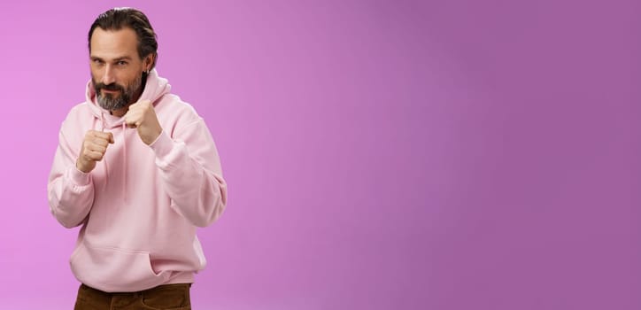 Sassy confident good-looking mature caucasian bearded man in pink hoodie teaching son how stand back fight defend himself raise fists ready boxing smiling daring, stay good shape, purple background.